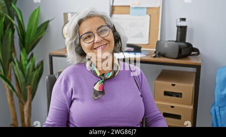 Eine lächelnde grauhaarige Frau mit Headset sitzt in einem modernen Bürobereich und strahlt Professionalität und Wärme aus. Stockfoto