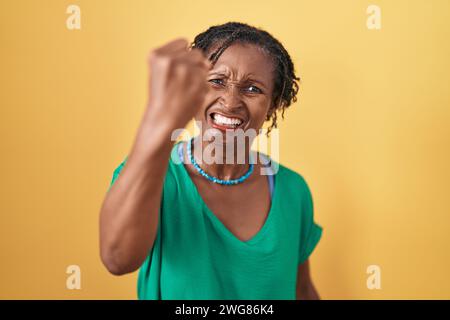Afrikanische Frau mit Dreadlocks, die über gelbem Hintergrund stehen, wütend und wütend mit der Faust, frustriert und wütend, während sie vor Wut brüllt. Rage und ag Stockfoto