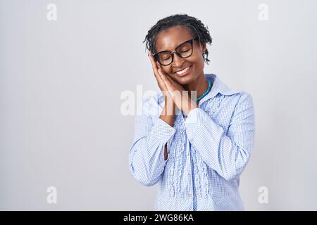 Afrikanische Frau mit Dreadlocks, die über weißem Hintergrund stehen und eine Brille tragen, die müde träumt und mit Händen zusammen posiert, während sie mit ihnen lächelt Stockfoto