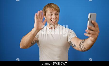 Blonder, bärtiger Mann in lässiger Kleidung, der ein Selfie macht und draußen vor blauem Hintergrund winkt. Stockfoto