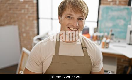 Porträt eines lächelnden kaukasischen Mannes mit blondem Bart, der eine Schürze trägt, in einem Kunststudio. Stockfoto