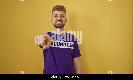 Ein hübscher junger Mann mit blauen Augen und Bart, der ein violettes „Freiwilliger“-T-Shirt trägt, zeigt und lächelt gegen eine gelbe Wand. Stockfoto