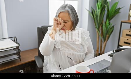 Eine gestresste Seniorin in einem Büro reibt sich die Augen, leidet an Müdigkeit oder Kopfschmerzen am Arbeitsplatz. Stockfoto