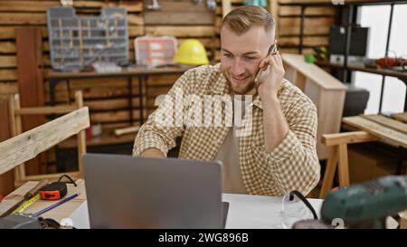 Ein junger kaukasier mit Bart arbeitet an seinem Laptop in einer Tischlerei, während er telefoniert. Stockfoto