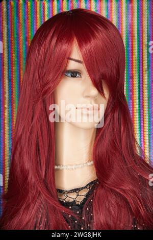 Plastikpuppe für Frauen mit hellen langen roten Haaren und schwarzer Spitze, die auf verschwommenem bunten Perlen-Hintergrund posiert. Ringlicht in ihren Augen. Stockfoto