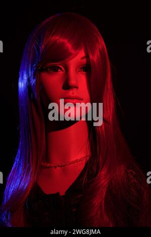 Plastikpuppe mit hellen langen roten Haaren, die auf schwarzem Hintergrund mit roten und blauen seitlichen Lichteffekten posiert Stockfoto