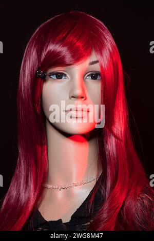Plastikpuppe mit hellen langen roten Haaren, die auf schwarzem Hintergrund mit rotem seitlichem Lichteffekt posiert Stockfoto
