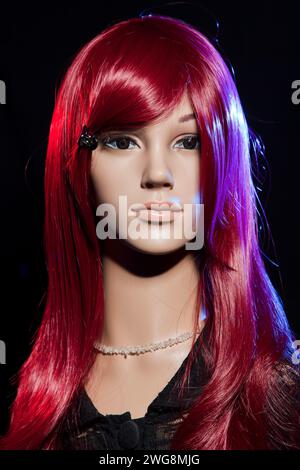 Plastikpuppe mit hellen langen roten Haaren, die auf schwarzem Hintergrund mit roten und blauen seitlichen Lichteffekten posiert Stockfoto