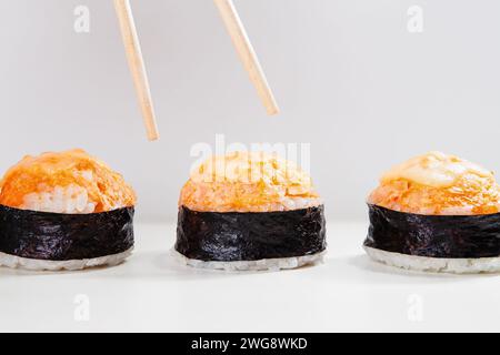 Ein Hauch von Essstäbchen, die positioniert werden, um ein Stück aus einem Trio von scharfen gebackenen Sushi-Nigiri oder Lachsgunkans auf weißem Hintergrund auszuwählen Stockfoto