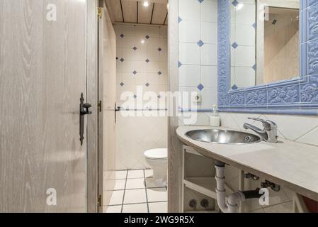 Ein kleines Badezimmer mit Porzellan und Holzwaschbecken unter einem blauen Porzellan gerahmten Wandspiegel und separater Toilette mit Tür Stockfoto