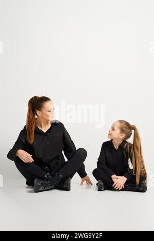 Mutter und Tochter sitzen auf weißem Hintergrund. Sie sind in stilvoller schwarzer Kleidung gekleidet und bieten einen modernen und legeren Look Stockfoto