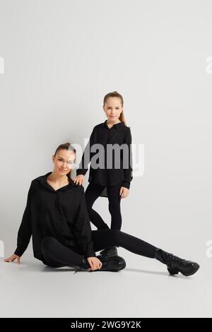 Mutter sitzt auf dem Boden und dehnt ein Bein zur Seite, und ihre Tochter steht hinter ihr und legt die Hand auf ihre Schulter. Frauen tragen ähnlich Stockfoto