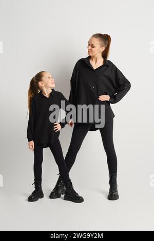 Eine moderne Familie in komplett schwarzem Outfit. Die Mutter und ihre Tochter tragen passende schwarze Stiefel, Hemden und enge Hosen und sehen sich gegenseitig an Stockfoto