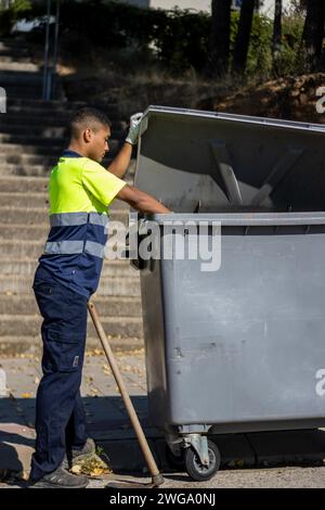 Vertikale Ansicht eines Gemeindearbeiters in Uniform, der einen Sack Müll in einen großen Müllcontainer wirft, während er den Deckel hochhält Stockfoto