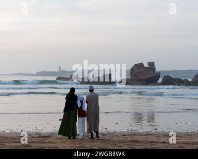 Die Familie steht am Meer und beobachtet, wie die Wellen über einer Felsformation mit der Insel dahinter bei Sonnenuntergang in Essaouira, Marokko, am 3. Februar 2024 hereinbrechen Stockfoto