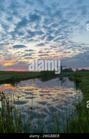 Eine typische niederländische Windmühle unter einer gesprenkelten Wolkendecke während des Sonnenaufgangs. Die gesamte Szene spiegelt sich im ruhigen Wasser des angrenzenden Grabens wider. Stockfoto