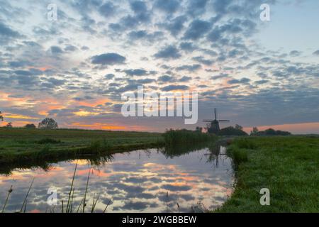 Eine typische niederländische Windmühle unter einer gesprenkelten Wolkendecke während des Sonnenaufgangs. Die gesamte Szene spiegelt sich im ruhigen Wasser des angrenzenden Grabens wider. Stockfoto