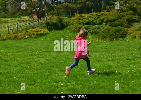 Ein entzückendes Kind in einer roten Jacke läuft durch eine grüne Wiese Stockfoto