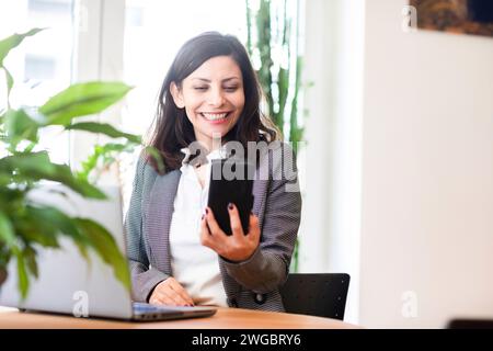 Lächelnde Geschäftsfrau, die an einem Schreibtisch vor ihrem Laptop sitzt und über ihr Mobiltelefon spricht Stockfoto