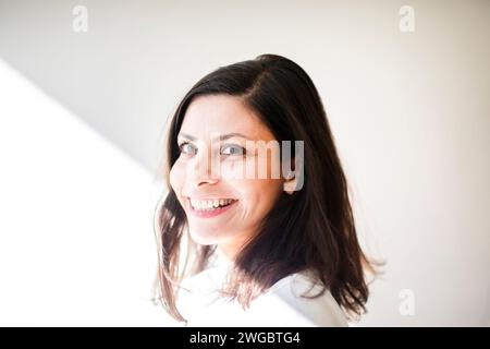 Porträt einer lächelnden Frau, die im Sonnenlicht an einer weißen Wand steht Stockfoto