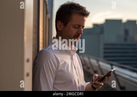 Ein blonder Mann Mitte dreißig mit einem dreitägigen Bart in weißem Hemd steht mit dem Rücken zur Wand auf der Terrasse eines Hochhauses A Stockfoto