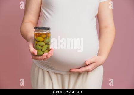 Schwangere Frau, die ein Glas Oliven in der Hand hält, rosafarbener Hintergrund. Schwangerschaftsbegriff und richtige Ernährung Stockfoto
