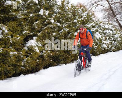 Ein fröhlicher Kerl in Sonnenbrille und orangefarbener Jacke fährt mit dem Fahrrad durch frisch gefallenen Schnee. Gesunder und aktiver Lebensstil im Freien im Winter Stockfoto