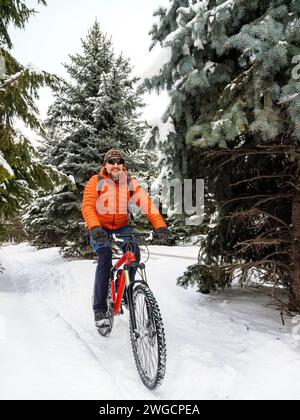 Ein Typ fährt mit dem Fahrrad in einem Winterpark. Ein Radfahrer in einer orangen Jacke fährt freudig auf einem verschneiten Pfad. Aktiver Lebensstil im Winter Stockfoto