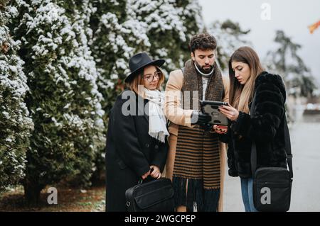 Eine Gruppe stilvoller Freunde, die an einem schneebedeckten Tag ein Tablet verwenden, winterliche Mode und Technologie. Stockfoto