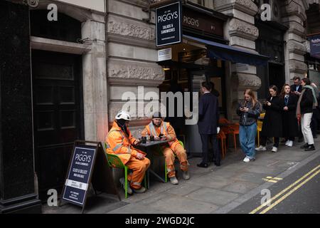 Handwerker sitzen draußen an einem Sandwich-Bartisch und überprüfen ihre Smartphones, während sie mittags einen Kaffee trinken, London, England, Großbritannien Stockfoto