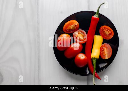 Anderthalb heiße Paprika auf einem schwarzen runden Teller. Kirschtomaten. Heller Hintergrund. Whiteboards. Gewürze in der Küche. Stockfoto