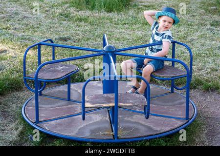 Ein Kinderspaß dreht sich auf dem Spielplatz und ein süßer Junge sitzt und ruht sich darauf aus. Stockfoto