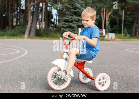 Ein kleiner Junge ohne Helm lernt, in einem Stadtpark ein Dreirad zu fahren. Stockfoto