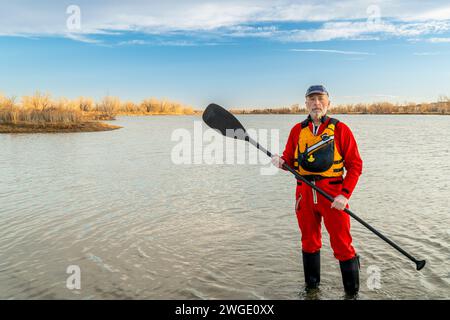 Umweltportrait eines männlichen Paddlers in einem Trockenanzug und einer Rettungsweste, die ein Stehpaddel an einem Seeufer hält Stockfoto