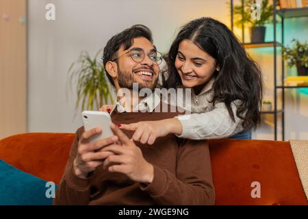 Lächelnde arabische Frau, die mit einem Freund spricht, der das Smartphone durchzieht und zu Hause auf dem Sofa sitzt. Multiethnisches junges Paar, das zusammen ein Mobiltelefon benutzt, um online Lieferung in Wohnzimmerwohnung zu bestellen. Stockfoto