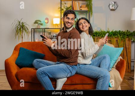 Seitenansicht eines fröhlichen, abwechslungsreichen Paares in legeren Kleidern, das sich hinten an hinten mit Smartphones auf dem Sofa im Wohnzimmer zu Hause sitzend. Junge Familie, die sich unterhalten, während sie ihr Handy auf der Couch in der Wohnung durchsucht. Stockfoto