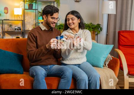 Glückliches junges Paar beim Online-Shopping, indem es die Kreditkartennummer auf dem Smartphone eingibt, während es zu Hause auf dem Sofa sitzt. Lächelnde Familie ist mit einer guten Aktion für Waren in der Wohnung zufrieden Stockfoto