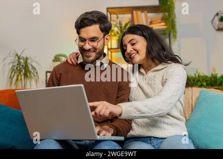 Fröhliches junges, abwechslungsreiches Paar in legeren Kleidern, während es im Wohnzimmer zu Hause mit Laptop auf Sofa diskutiert. Glückliche indische Familienplanung zusammen zeigt, wie man online Tickets in der Wohnung Bucht Stockfoto