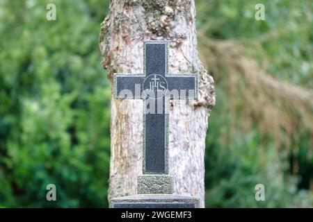 Das christliche IHS-Monogramm auf einem Steinkreuz auf einem Friedhof vor einem Baum in einem verschwommenen Hintergrund Stockfoto