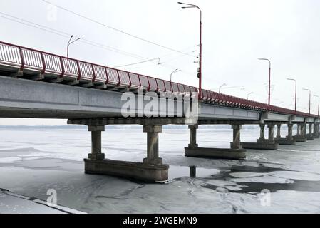 Straßenbrücke auf dem Winterfluss. Große Betonbrücke mit roten Geländern und Lampenpfosten. Der Fluss ist mit Eis bedeckt. Stockfoto