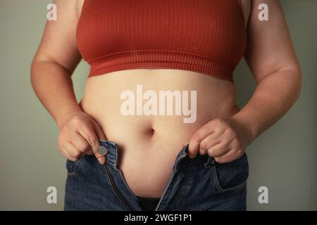 Frauen Körperfettbauch. Übergewichtige Frauenhand mit übermäßigem Bauchfett. Eine fette Frau, die versucht, ihre Jeanshose zu ziehen. Frauengesundheit. Stockfoto