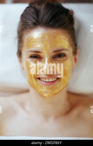 Gesundheitszeit. Obere Ansicht der lächelnden modernen 40-jährigen Frau in Massagekabine mit goldener Kosmetikmaske auf dem Gesicht, das auf Massagetisch liegt. Stockfoto