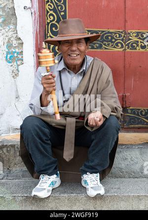 Ein Mann dreht ein handgehaltenes tibetisches Gebetsrad, während er auf Steintreppen auf dem Barkhor Square in Lhasa sitzt. Stockfoto