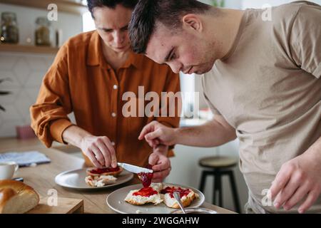 Junger Mann mit Down-Syndrom, der das Frühstück mit seiner Mutter zuhause zubereitet. Morgenroutine für Mann mit Down-Syndrom. Stockfoto