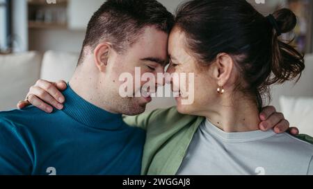 Porträt eines jungen Mannes mit Down-Syndrom mit seiner Mutter zu Hause, in der Hand, mit der Stirn berührt. Das Konzept der Liebe und der Erziehung behinderter Kinder Stockfoto