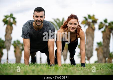 Fröhlich zufriedene und athletische Männer und Frauen sitzen im Gras bei Sportbekleidung Training draußen. Schönes, junges, erwachsenes Paar, das sich in der Gegend ausdehnt Stockfoto
