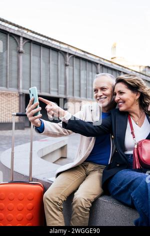 Fröhliches Touristenpaar, das zusammen reist, um Selfie zu machen und draußen mit Gepäck zu sitzen. Liebevoller Ehemann und Ehefrau machen Fotos Stockfoto