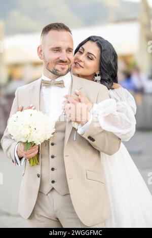 Hochzeitsporträt lächelnder Jungvermählter. Ein stilvoller Bräutigam in einem beigefarbenen Anzug und eine süße Brünette in einem weißen Kleid sind zärtlich im Park. Stockfoto