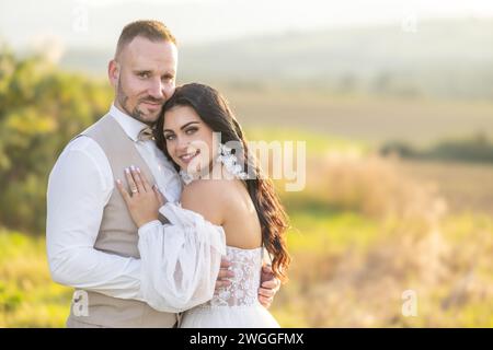 Hochzeitsporträt lächelnder Jungvermählter. Ein stilvoller Bräutigam in einem beigefarbenen Anzug und eine süße Brünette in einem weißen Kleid sind zärtlich im Park. Stockfoto