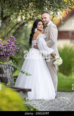 Hochzeitsporträt lächelnder Jungvermählter. Ein stilvoller Bräutigam in einem beigefarbenen Anzug und eine süße Brünette in einem weißen Kleid sind zärtlich im Park. Stockfoto